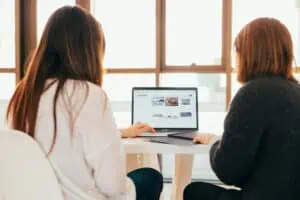 Two people sit at a table, viewing a laptop screen displaying a website.